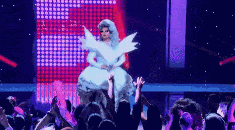a drag queen in a white dress with wings is being lifted by a crowd of people .