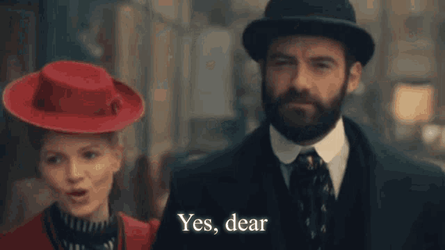 a man in a suit and tie is standing next to a woman in a red hat and saying yes , dear .