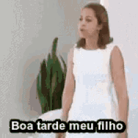 a woman in a white dress is standing in front of a plant and saying `` boa tarde meu filho '' .