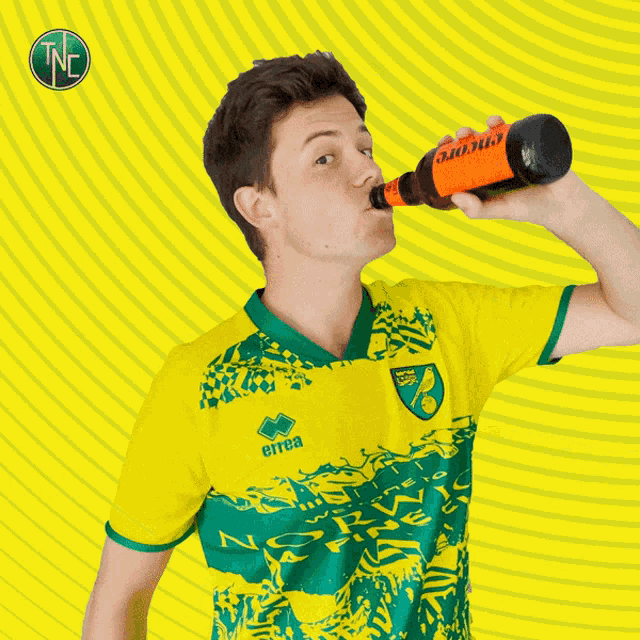 a man in a yellow and green errea shirt is holding a bottle of coke