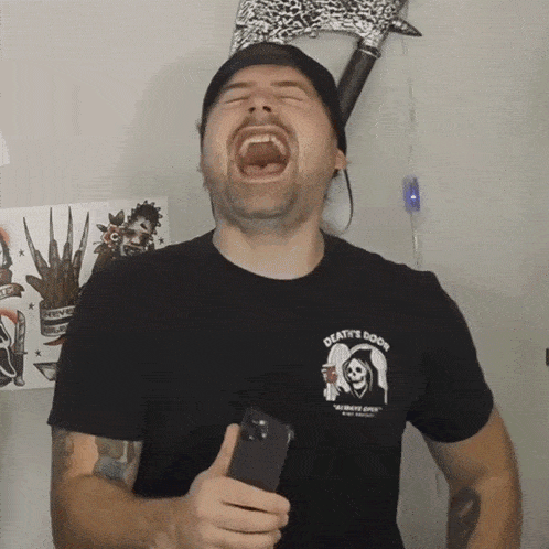 a man wearing a death 's door t-shirt laughs while holding a cell phone
