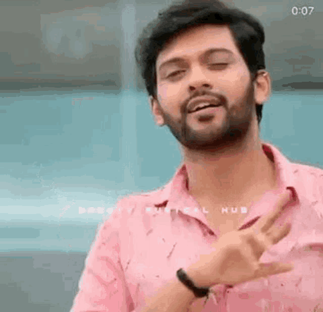 a man with a beard is wearing a pink shirt and making a gesture with his hands .