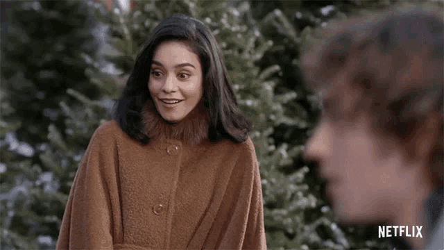 a woman in a brown coat is standing next to a man in front of a christmas tree and smiling .