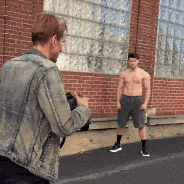 a man in a denim jacket takes a picture of a shirtless man in shorts