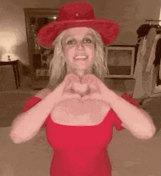 britney spears is wearing a red dress and a red hat and making a heart shape with her hands .