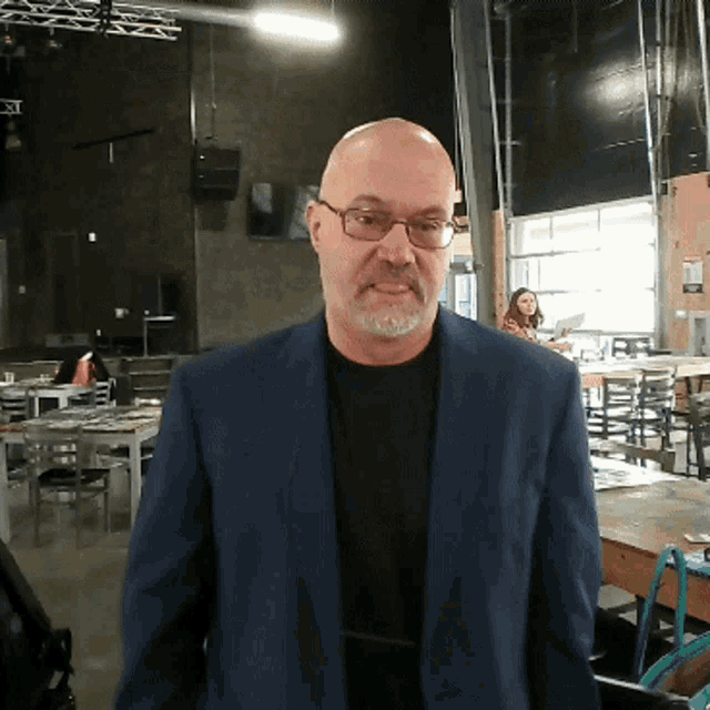 a bald man wearing glasses and a blue jacket is standing in a restaurant