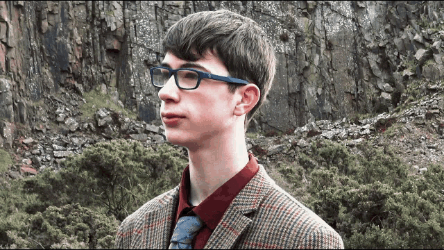 a young man wearing glasses and a plaid jacket stands in front of a mountain