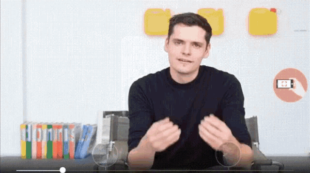 a man in a black shirt is talking in front of a white wall