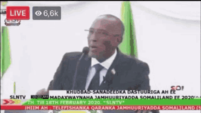 a man in a suit and tie is speaking into a microphone on a screen that says live