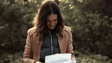 a woman in a brown jacket is reading a piece of paper with the bbc logo in the background