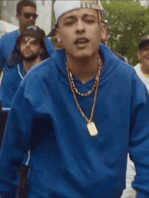 a man wearing a blue hoodie and a gold necklace is surrounded by other men