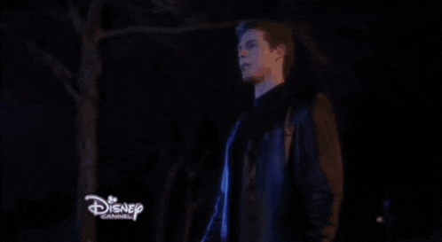 a man is standing in a dark room with his arms outstretched in front of a disney channel logo .