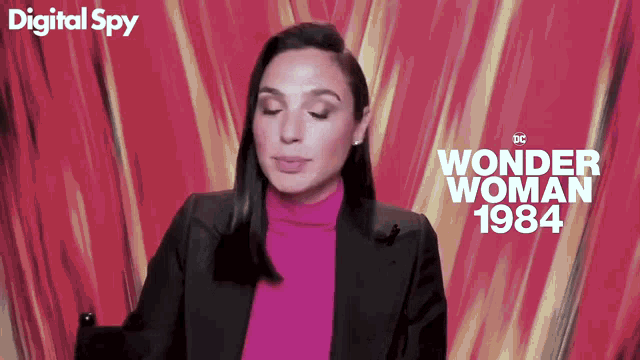 a woman in a pink turtleneck and black jacket is sitting in front of a digital spy wonder woman 1984 poster