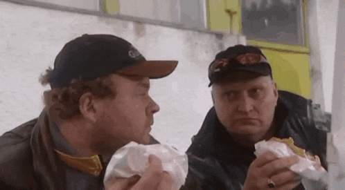 two men are eating hamburgers and smoking a cigarette .