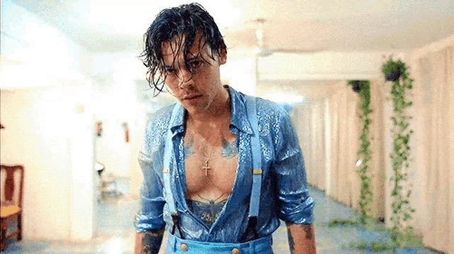 harry styles is wearing a blue shirt and suspenders and is standing in a hallway .