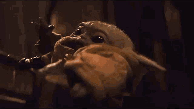 a baby yoda is sitting on a chair in a dark room and waving at the camera .