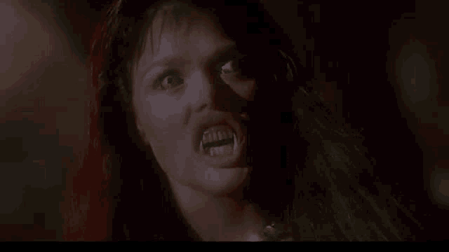 a woman with long red hair and vampire teeth is looking at the camera in a dark room .