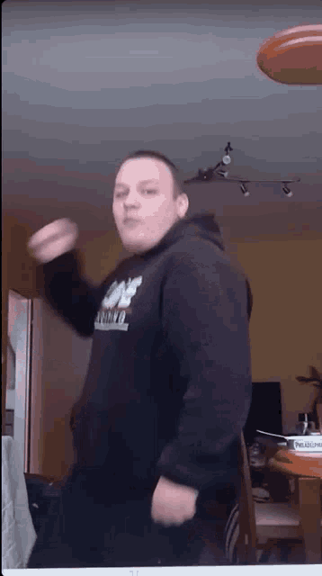 a man in a black hoodie is dancing in a living room with a frisbee in the background .