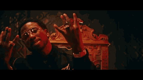 a man wearing glasses and a watch is sitting on a throne with his hands in the air .