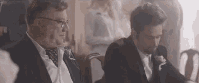 two men in suits are sitting next to each other in a room and talking .