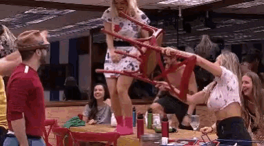 a group of people are standing around a table and a woman is holding a red chair .