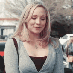 a woman wearing a grey cardigan and a necklace is standing in a parking lot .