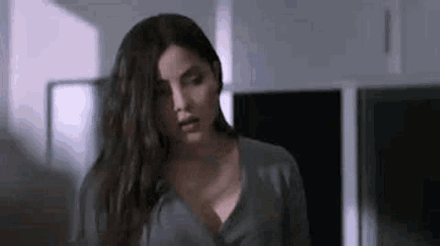 a woman is standing in a kitchen wearing a gray sweater and looking at the camera .