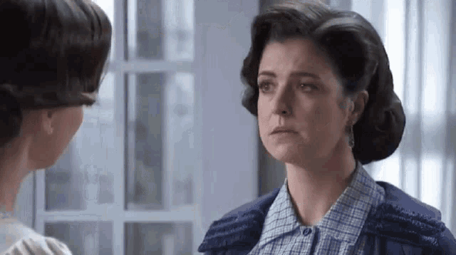 two women are standing next to each other in front of a window and talking to each other .