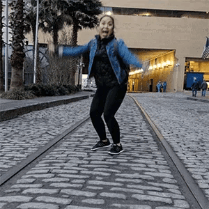 a woman in a denim jacket is jumping in the air on a cobblestone street