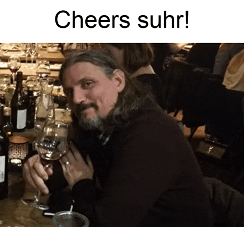 a man with long hair and a beard is sitting at a table holding a glass of wine with the words cheers suhr below him