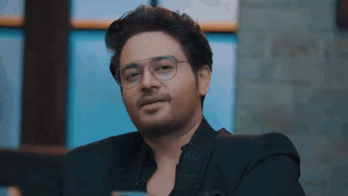 a man wearing glasses and a black suit is sitting in front of a window and smiling .