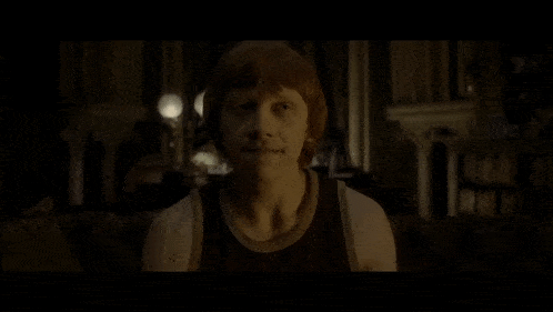 a young man with red hair and a black tank top smiles in a dark room