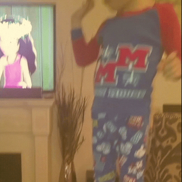 a boy wearing a blue shirt with the letter m on it stands in front of a television