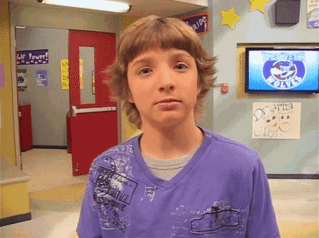 a boy in a purple shirt stands in front of a wall with a sign that says drama club on it
