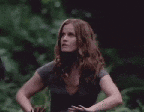 a woman with long red hair is standing in the woods with her hands on her hips