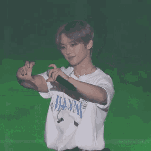 a young man in a white t-shirt is holding a microphone and making a heart shape with his hands .