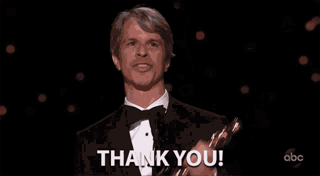 a man in a tuxedo is holding an oscar trophy and saying `` thank you '' .