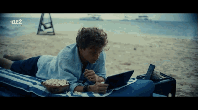 a man laying on a beach with a bowl of popcorn and a tablet that says tele2 on the bottom