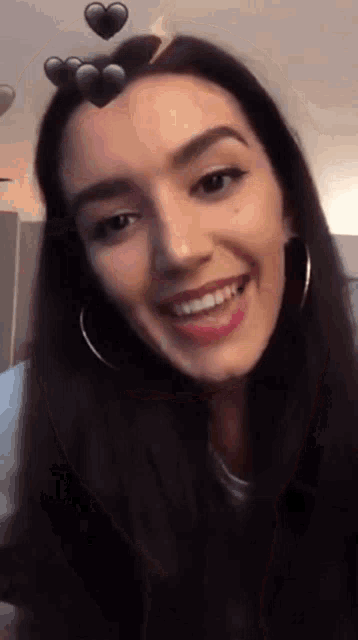 a woman with long dark hair and hoop earrings is smiling with hearts floating around her head .