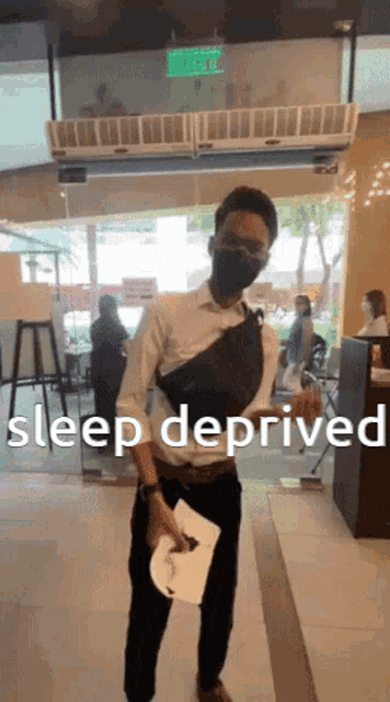 a man wearing a mask holds a piece of paper in front of a sign that says " sleep deprived "