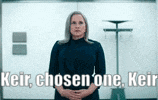 a woman in a blue dress is standing in front of a door with the words keir chosen one keir written above her .