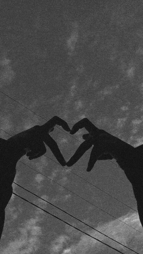 a couple of hands making a heart shape with their fingers against a cloudy sky .