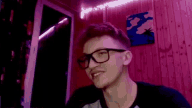 a young man wearing glasses and a black shirt is sitting in front of a pink wall .