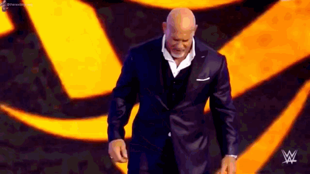 a bald man in a suit is walking on a stage in front of a yellow and orange background .