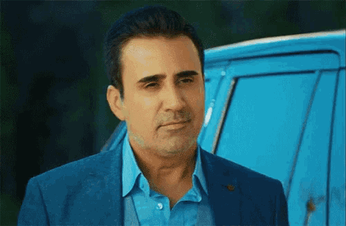 a man in a suit and blue shirt is standing in front of a blue car