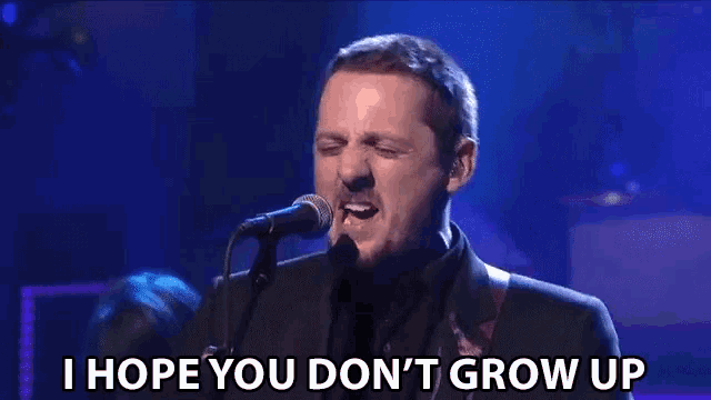 a man singing into a microphone with the words " i hope you don 't grow up " behind him