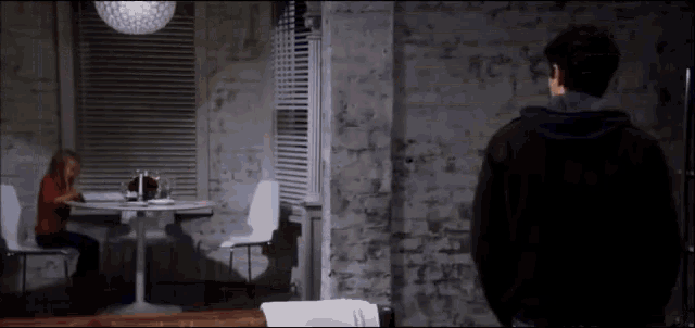 a man in a black hoodie stands in front of a woman sitting at a table in a dark room