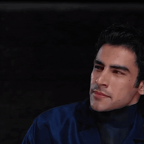 a man in a blue jacket and black turtleneck is looking at the camera with a serious look on his face .