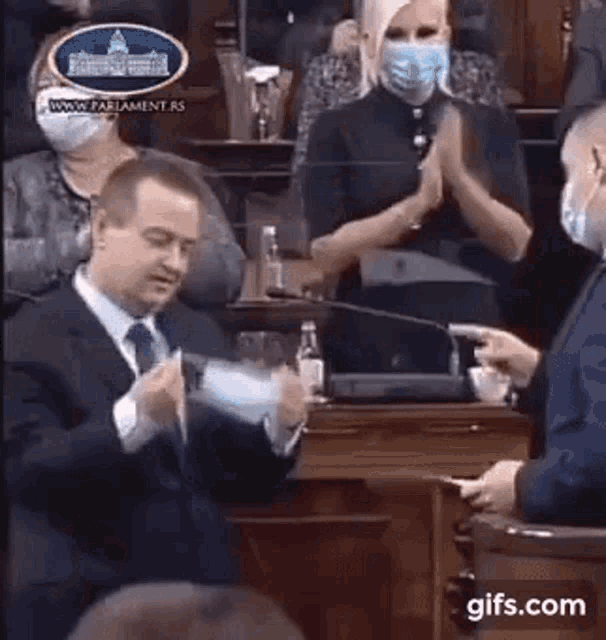 a man in a suit and tie is standing in front of a microphone while a woman in a mask applauds .