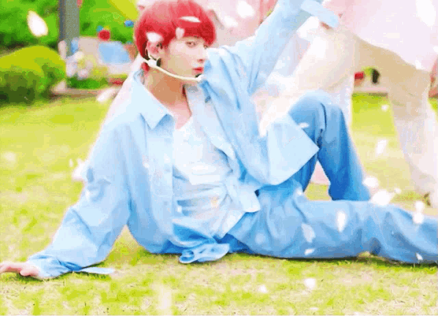 a man with red hair is sitting on the grass wearing a blue shirt and blue pants .